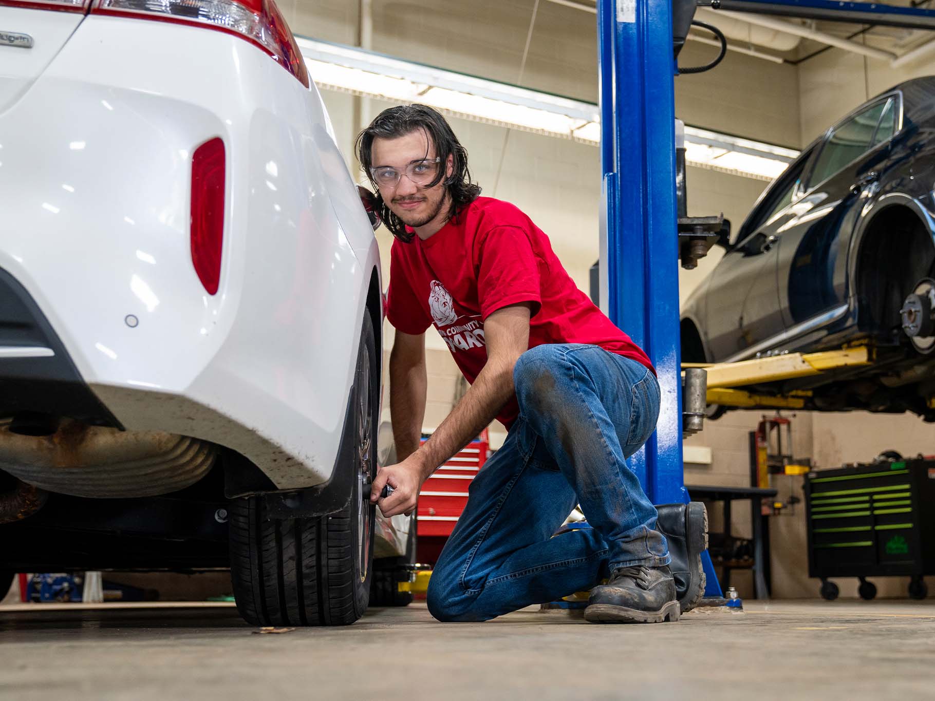 An automotive student wearing a red T‑shirt and safety glasses kneels beside a white car in a repair shop, tightening a wheel lug nut, with vehicle lifts and other cars in the background.