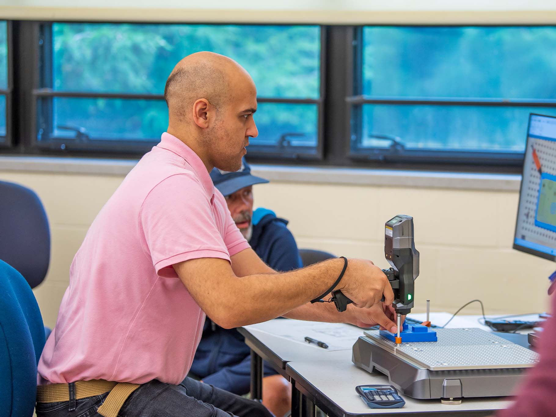 A man in a pink shirt operates a small precision manufacturing or testing device at a workbench in a classroom or lab setting, with another person seated behind him and a computer monitor displaying technical information nearby.